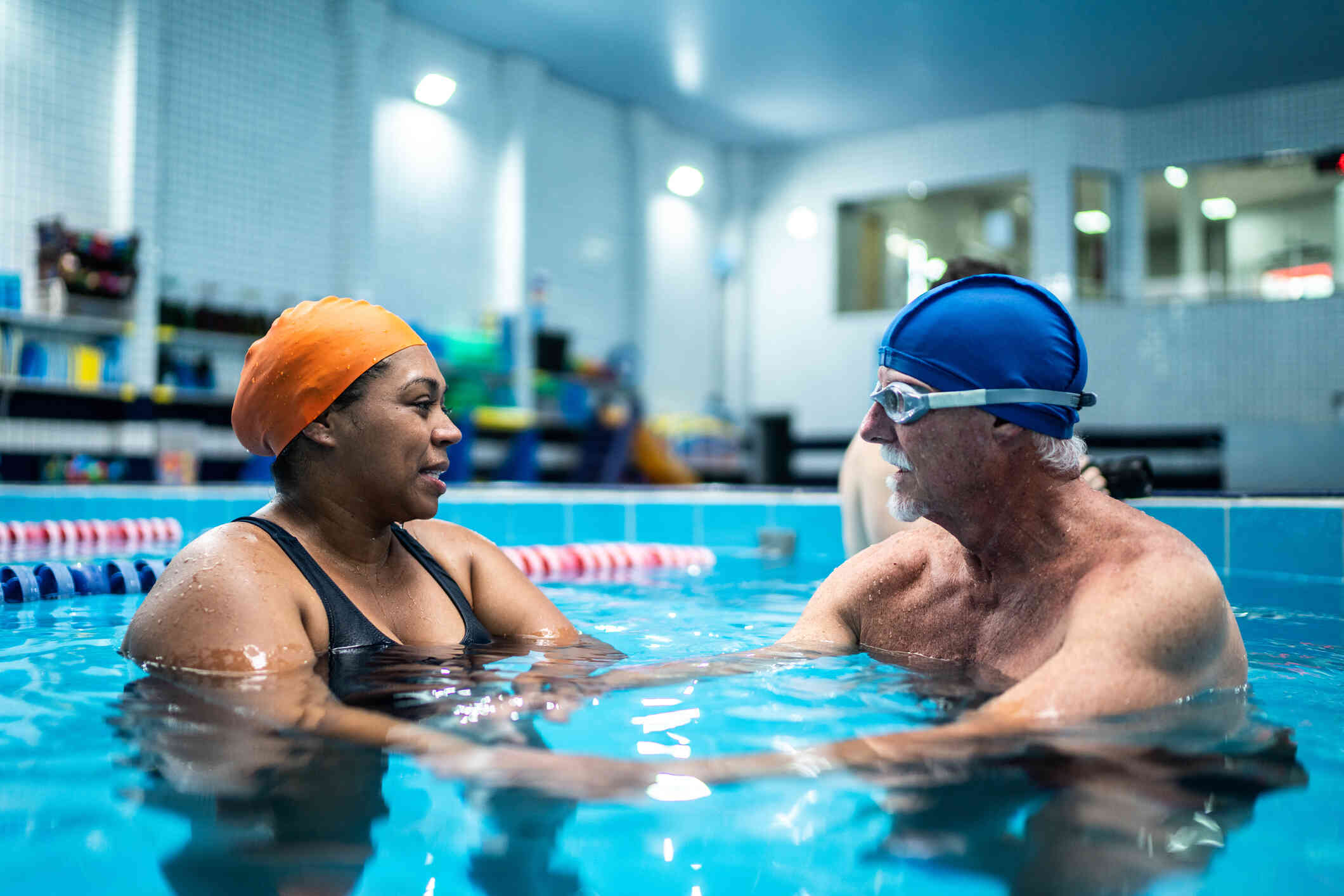 A woman and an elderly man in a swim cap talk to each other while in an indoor pool.